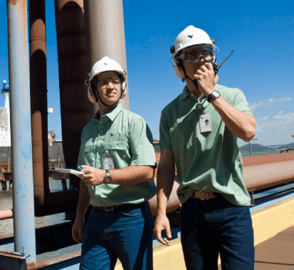 Two Vale employees are in an outdoor operation area. Both are wearing helmets and goggles, and one of them is talking on a radio transmitter.