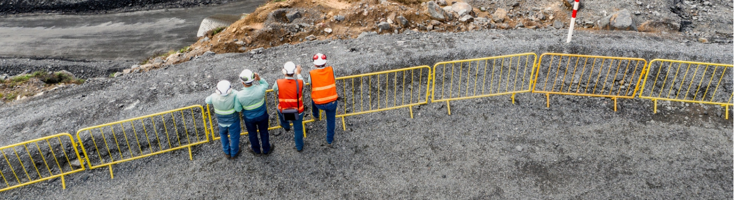 A top-down shot of four people observing an operation area near the grids. Two of them are wearing jeans, orange vests with fluorescent details and helmets. The other two are wearing jeans, green Vale shirts with some fluorescent details and helmets.