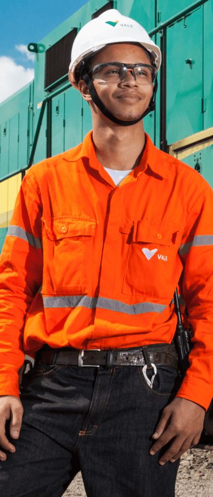 A man standing next to a green, yellow and gray train. He is wearing orange uniform with Vale logo, goggles, and a white helmet also with the company logo.