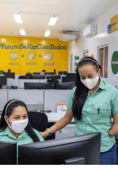 Two Vale female employees are side by side in an office. One is sitting in front of a computer and the other is standing. Both wear light green uniform shirts and white face masks