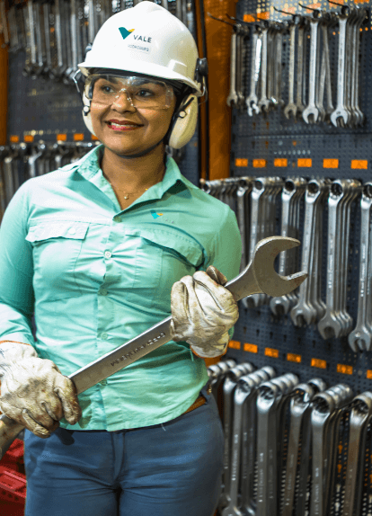 A woman standing, holding a steel tool in a space full of other tools. She is wearing light green uniform, gloves, goggles, ear muffs and a white helmet with Vale logo.