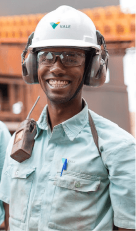 Vale employee smiling in an operational area. He is wearing light green shirt with Vale logo, radio communicator fixed to the shoulder, goggles, ear muffs and a white helmet with the company logo.