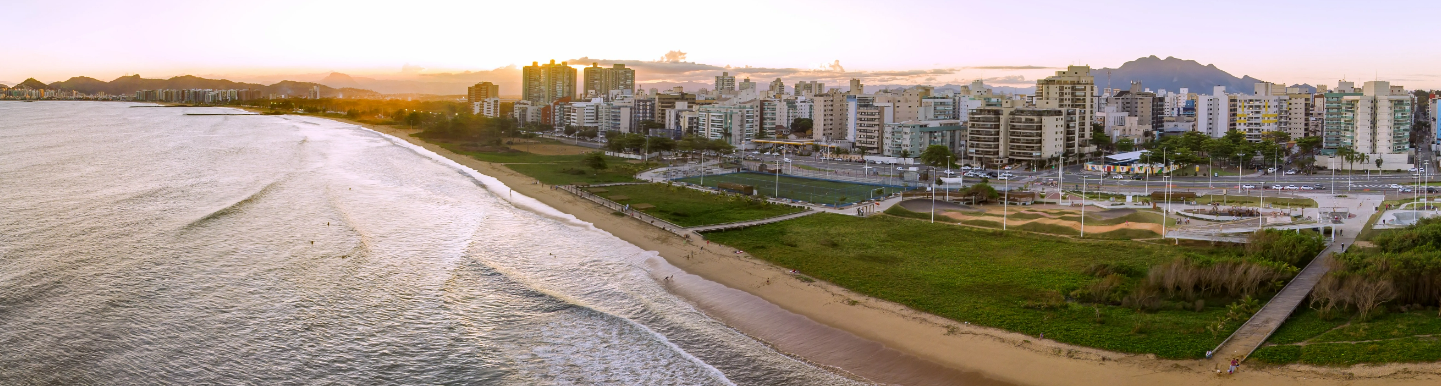 Imagem aérea da praia de Camburi. Ao fundo há uma área gramada e diversos prédios.