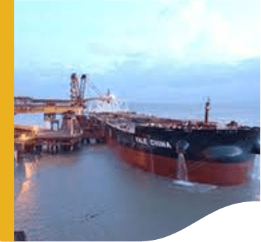 Image of a large cargo ship docked at the port. On the left side, there are several crane-like machines connected to the ship.