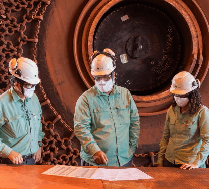 Two men and a woman stand side by side working in an operational space. They are wearing long-sleeved shirts in a light shade of green, helmets, goggles, and protective masks. In the background is a wheel of a large vehicle.