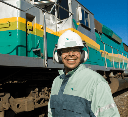 Empregado Vale sorrindo. Ele usa capacete e óculos de proteção. Ao fundo há um trem Vale, nas cores verde e amarela.