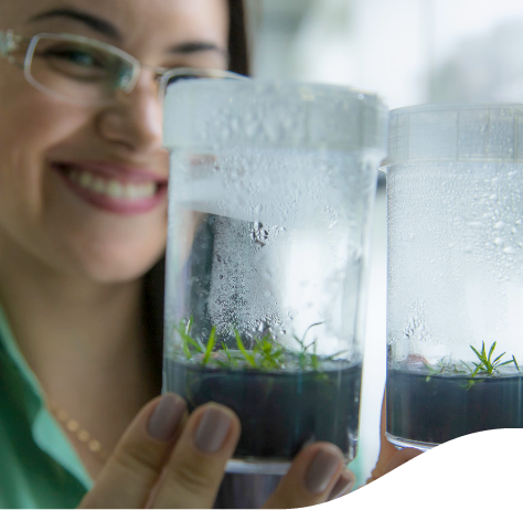 In the foreground, there are two vases with germinating seeds, in the background a woman - holding the vases - smiles. She wears glasses and a small chain.