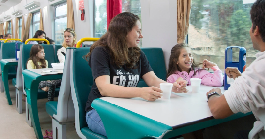 Uma família, composta por uma mulher, um homem e uma menina, estão sentados ao redor de uma mesa que contém dois copos em cima. Ao fundo é possível ver outras pessoas sentadas.