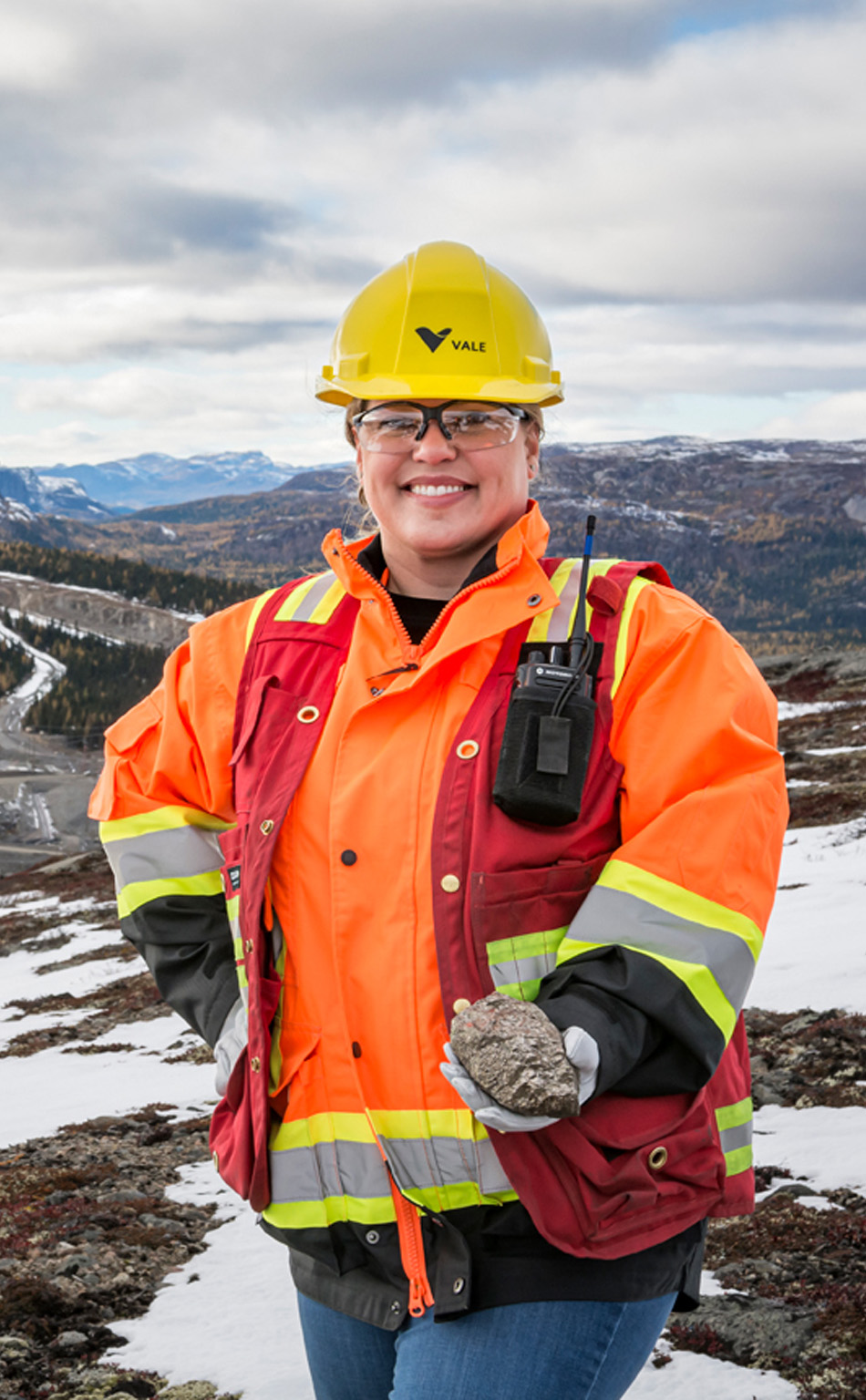 Funcionária da Vale sorrindo em paisagem verde. Ela veste uniforme verde
da vale, oculos, capacete e protetores auriculares