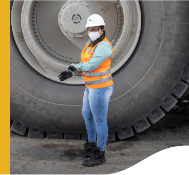 Photo of a woman wearing Vale uniform, green button-down shirt, orange vest with fluorescent illumination, a helmet, a mask, goggles, gloves and boots. In the background, there is a huge truck wheel, twice the height of the woman.