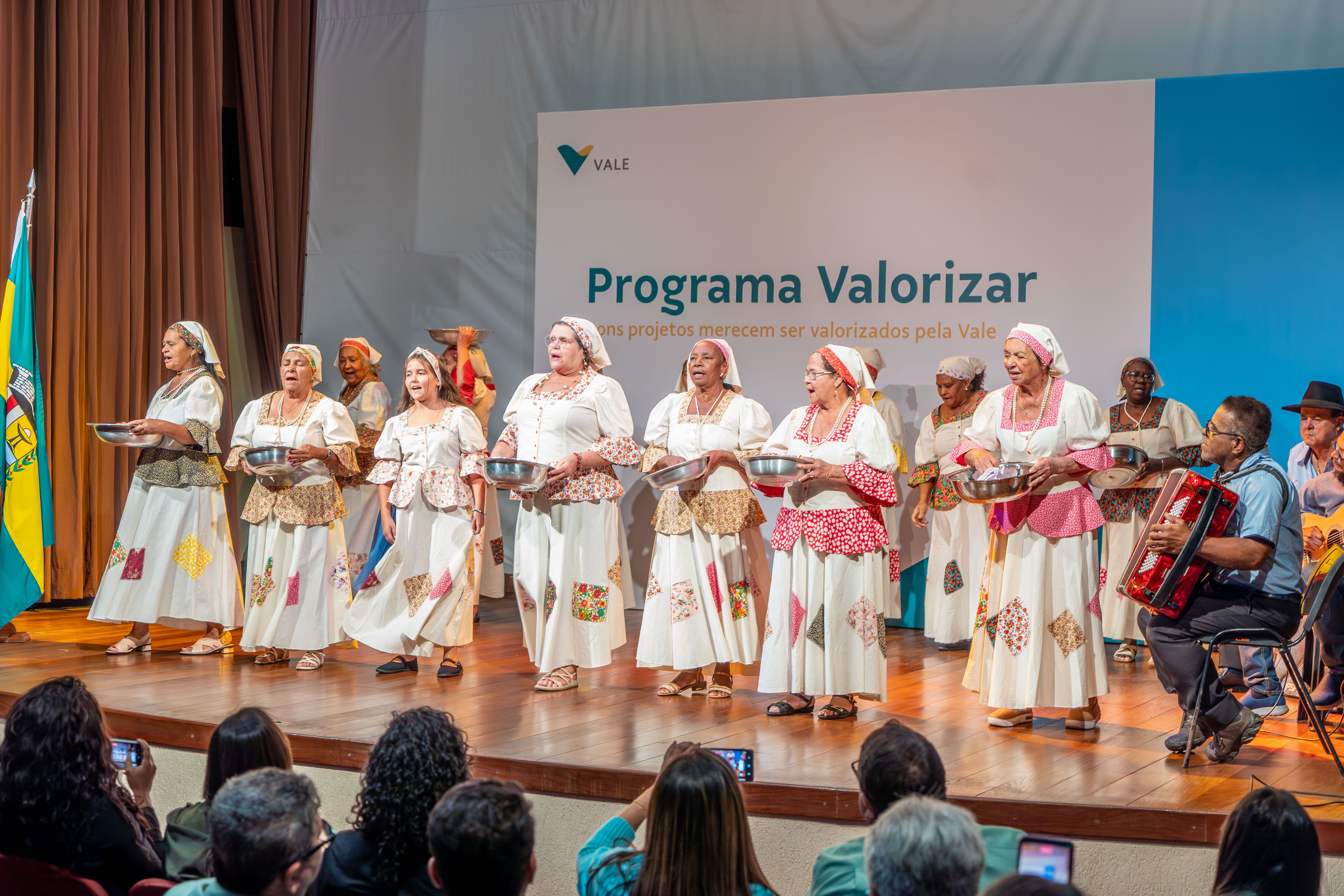 Diversas mulheres com vestidos iguais se apresentam em cima de um palco, elas seguram uma bacia nas mãos. Ao lado delas há um sanfoneiro.