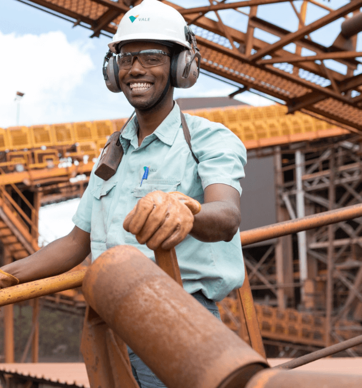 In an outdoor operation area, a Vale employee is smiling for the photo. He is wearing goggles, a helmet and gloves. On his shoulder, there is a radio transmitter fixed.