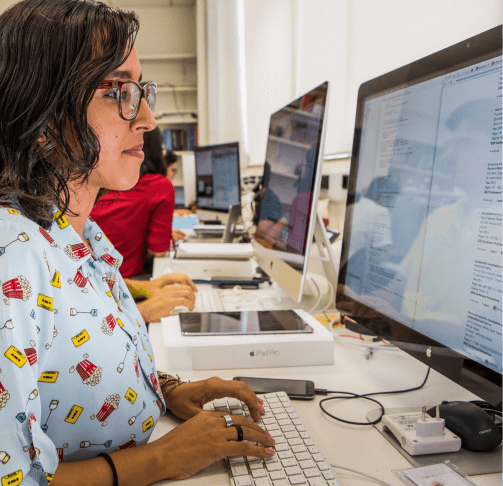 A woman in a printed blouse types on a computer. In the background you can see other people doing the same.