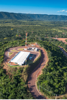 Foto vista de cima de uma área grande verde com vegetação e no meio uma estrutura de um galpão com postes em volta e um caminho asfaltado.