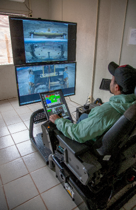 Um empregado está sentado em uma cadeira eletrônica, operando equipamentos remotos por meio de duas telas.