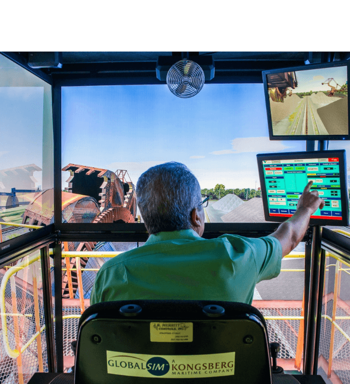 Empregado Vale, sentado de costas para foto, operando remotamente um equipamento. Ele está tocando em uma das duas telas que há em sua frente. Ao fundo, é possível ver um maquinário grande.