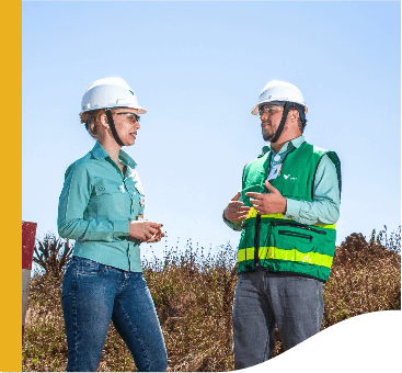 Dois empregados Vale, ambos de capacete de proteção, conversam em uma área aberta, com uma vegetação rasteira ao fundo.