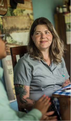 Uma empregada Vale está sentada, olhando para o lado e sorrindo. Ela tem cabelo loiro e usa um uniforme cinza