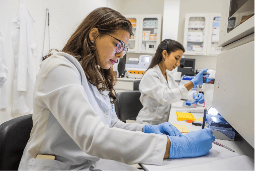 In a laboratory, two women in gloves and white lab coats work in concentration. One of them takes notes.