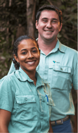 hoto taken from the waist up of two Vale employees – a man and a woman – smiling in an area with trees. The two wear light green shirts.