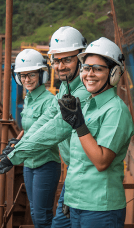 Três empregados – duas mulheres e um homem – parados lado a lado em uma estrutura de metal dentro de uma operação. Os três estão sorrindo e usam camisas em um tom de verde claro, óculos de proteção, protetores de ouvido e capacete branco com logotipo da Vale.