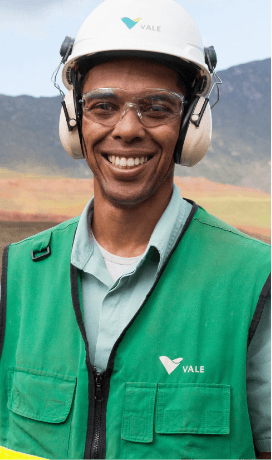 Homem sorrindo em um espaço operacional. Ele usa camisa verde clara e um colete em um tom de verde mais escuro com o logotipo da Vale em branco. óculos de proteção, protetores de ouvido e capacete branco com logotipo da Vale.