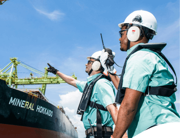 Dois empregados da Vale de frente para um navio em uma área operacional. Os dois usam camisa verde, equipamentos de proteção presos ao corpo, óculos de proteção, protetores de ouvido e capacete branco.