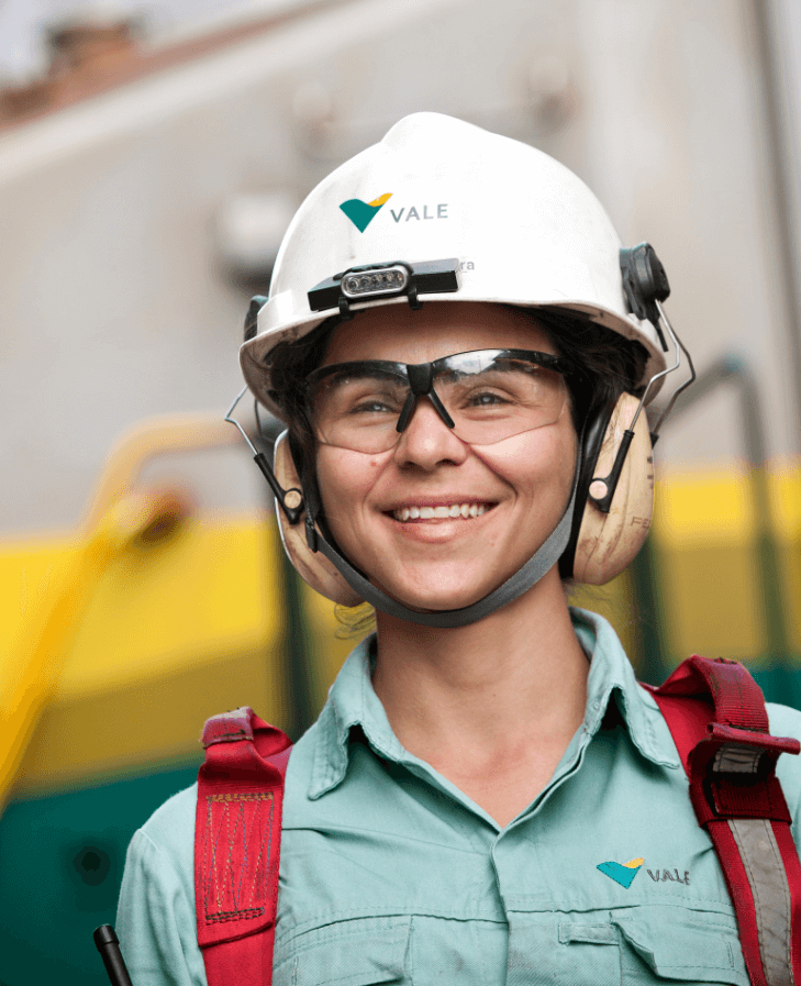 Mulher sorrindo em uma área operacional. Ela usa camisa verde clara com logotipo da Vale, equipamento de segurança vermelho preso aos ombros, óculos de proteção, protetores de ouvido e capacete branco com logotipo da Vale.