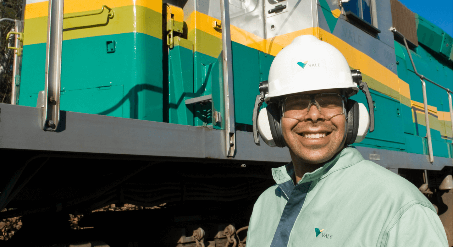 A man smiles for the photo. He is wearing a light green uniform, ear protectors, goggles, and a white helmet with the Vale logo. Behind him is a green and yellow locomotive.