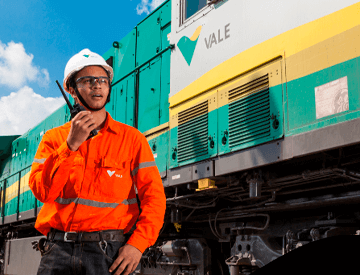 Homem parado ao lado de um trem verde, amarelo e cinza. Ele usa uniforme laranja com logotipo da Vale, óculos de proteção e capacete branco também com logotipo da empresa.