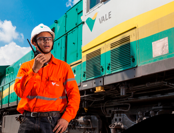 A man standing next to a green, yellow and gray train. He is wearing orange uniform with Vale logo, goggles, and a white helmet also with the company logo.