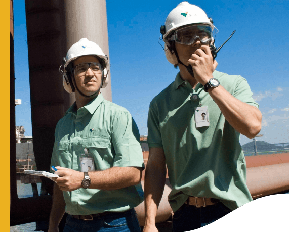 Foto de dois homens em uma parte alta de uma operação observando algum ponto. Um dos homens está segurando e falando em um rádio com uma mão, ele está usando camisa polo verde clara com logo da Vale, crachá, relógio, proteção nos ouvidos, capacete e óculos de proteção. Já o outro está segurando um caderno e uma caneta e está usando camisa de botões verde clara com logo da Vale, crachá, relógio, proteção nos ouvidos, capacete e óculos de proteção.