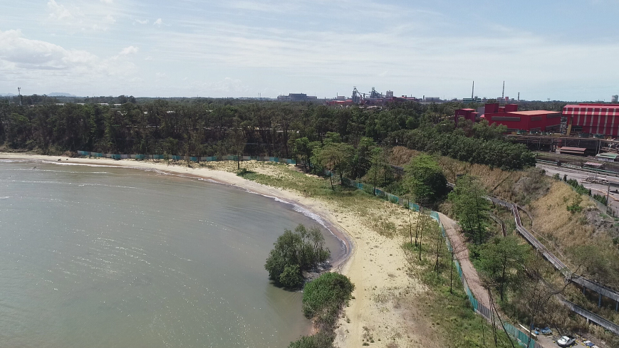 Água da praia em visão ampliada com vegetação viva, árvores e areia ao redor. Há também construções urbanas, carros e estradas ao fundo.