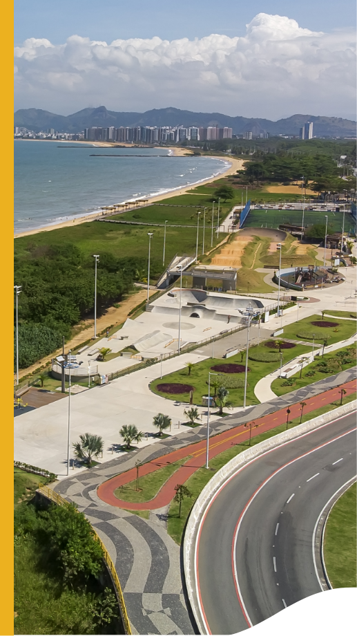 Vista área da cidade. Ao fundo, o mar, com areia e vegetação viva ao redor, cercada por construções urbanas, árvores e pessoas.