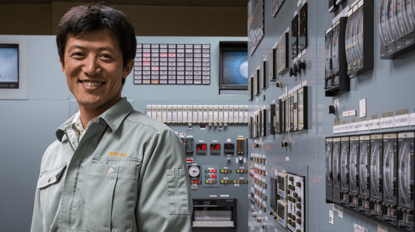 A Vale employee smiles for the photo. He is wearing a gray uniform. Behind him is a control room