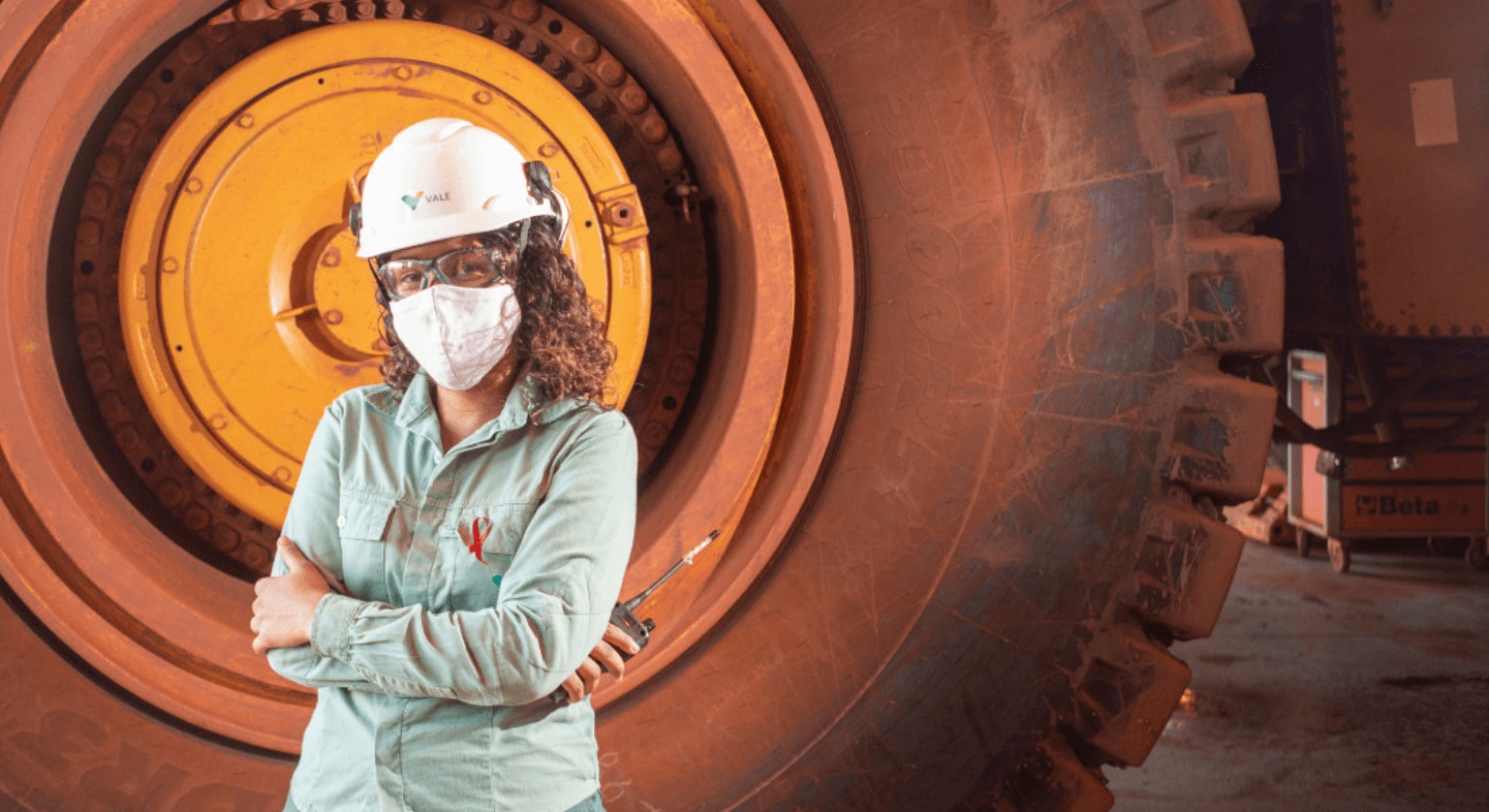 A woman stands with her arms crossed and holds a communication radio in one hand. She is wearing a light green uniform, helmet, goggles, and mask. In the background is a truck wheel.