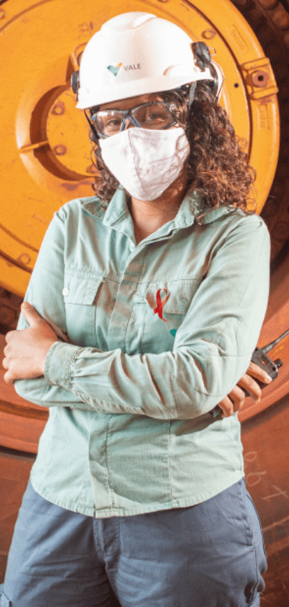 A woman stands with her arms crossed and holds a communication radio in one hand. She is wearing a light green uniform, helmet, goggles, and mask. In the background is a truck wheel.