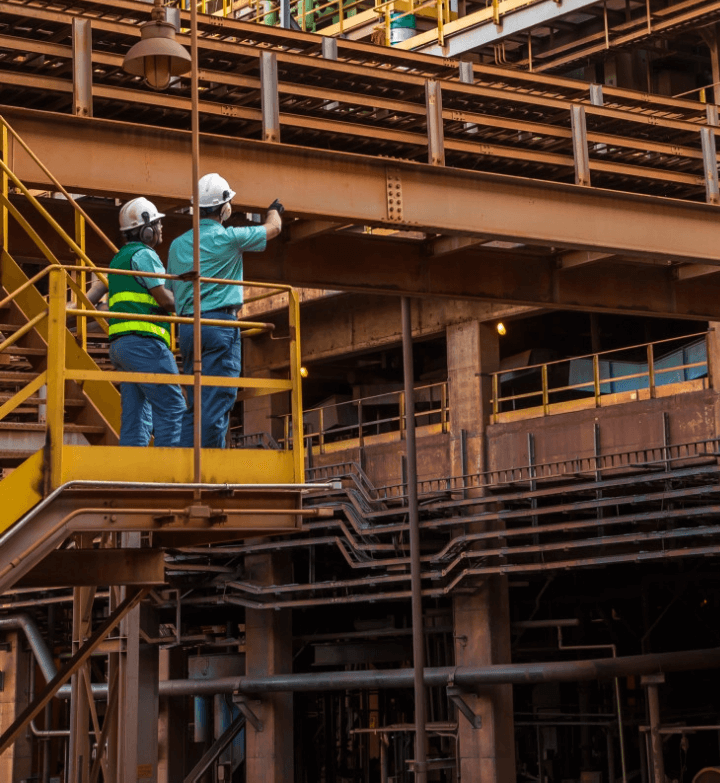 Two Vale employees stand under an iron structure and one of them points upwards. Both are wearing protective helmets.