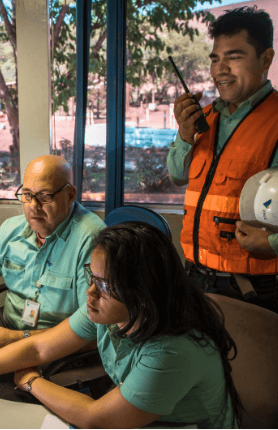 Two o Vale employees – a man and a woman – sitting side by side at an office table and looking straight ahead. The two wear light green shirts. Behind them, there is another employee, wearing a green shirt and an orange vest, in one hand, he holds a white helmet, and in the other, a radio communicator.