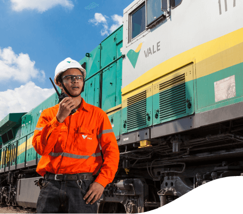 Foto de um homem na frente de um trem da Vale pintado de amarelo, verde e cinza. Ele está segurando um rádio e está usando camisa fluorescente laranja, capacete, óculos de proteção, cinto e calça jeans.