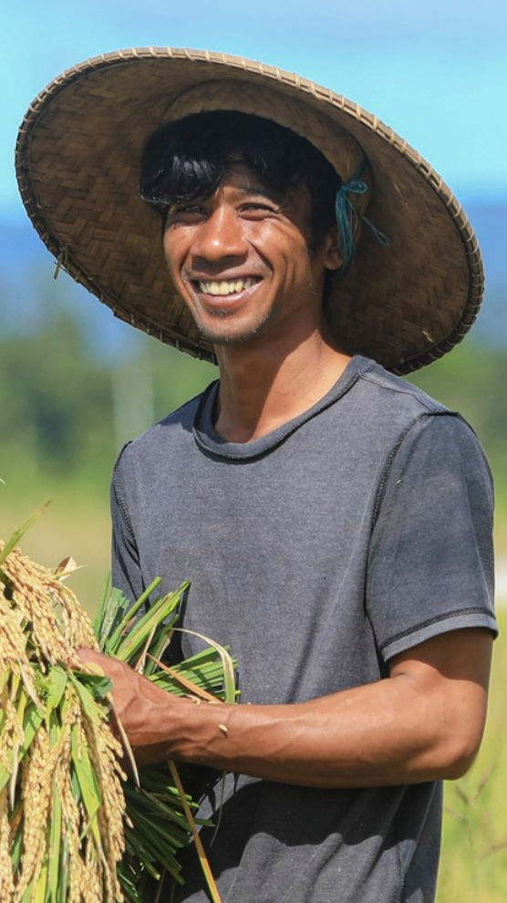 Seorang pria bertopi tersenyum untuk difoto. Di tangannya ia membawa beberapa tanaman.