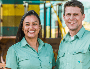 Foto tirada da cintura para cima de dois funcionários da Vale – um homem e uma mulher – sorrindo em um espaço de operações. Os dois usam camisas verdes em um tom claro.