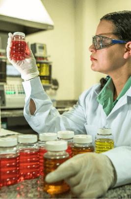 A researcher holds red liquid samples. She is wearing a white lab coat, gloves and goggles.