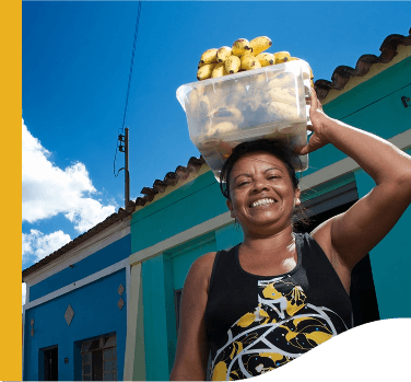 A woman smiles for a photo while holding a transparent bowl with bananas on her head. In the background are two blue houses of ancient architecture.