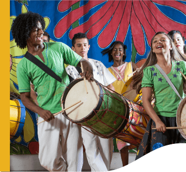 Young people play drums and smile, as if they were singing. At the back there is a fabric printed with the colors blue, red, yellow, and green.