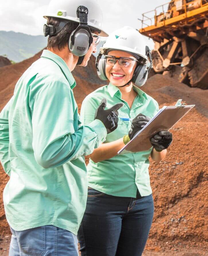 Dois funcionários da Vale – um homem e uma mulher – parados lado a lado em um espaço de operações com um grande veículo ao fundo. Os dois usam camisa verde, luvas, óculos de proteção, protetores de ouvido e um capacete branco. A mulher segura uma prancheta e sorri.