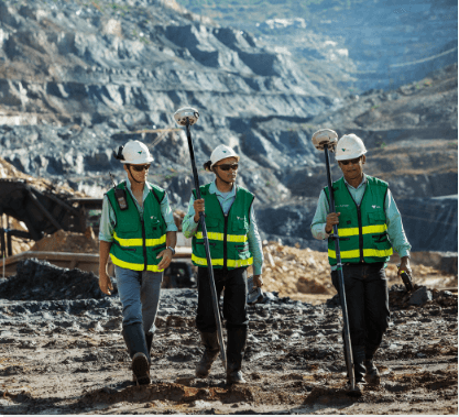 Photo from knee to head height of three men wearing Vale's light green shirt, green vest with yellow markings, helmet and goggles. They seem to be walking in an operation.