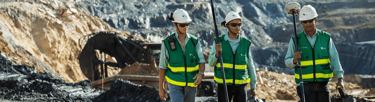 Foto da altura do joelho até a cabeça de três homens utilizando camisa verde clara da Vale, colete verde com sinalizações amarelas, capacete e óculos. Eles parecem estar andando em uma operação.