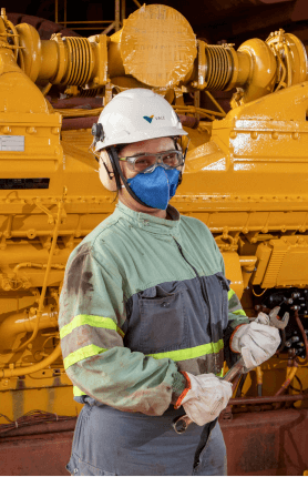 Photo from thigh to head of a woman wearing Vale's green and gray overalls, helmet, ear protection, goggles and mask, holding a tool with a machine in the background.