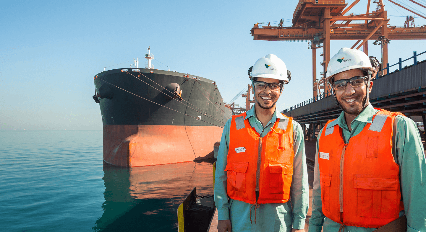 Dois homens em uma área de operação próximo ao mar estão sorrindo. Eles usam camisa em um tom de verde claro, colete laranja, capacete branco e óculos de proteção.